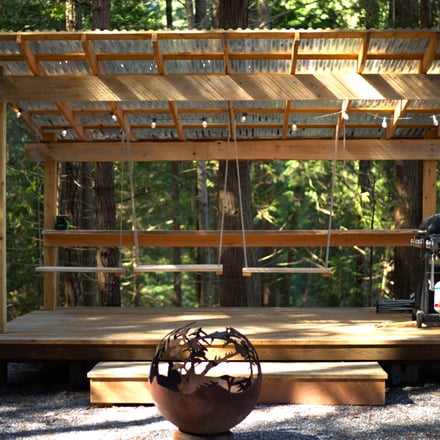 Wooden pergola with metal fire pit sphere in forest clearing, grill visible on right side