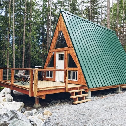 A-frame cabin with wood siding and green metal roof surrounded by tall evergreen trees and rocky ground.