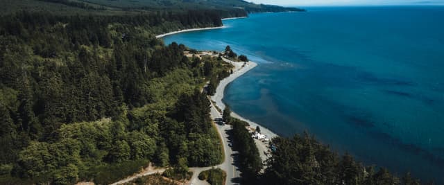 Aerial view of turquoise lake with rocky shoreline, dense coniferous forest, mountains, and winding road along the coast