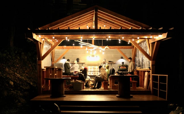Illuminated wooden pavilion at night with guests gathered around a central kitchen counter and cooking area